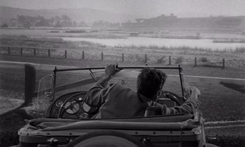 Movie still from “The Killing” (1956), directed by Stanley Kubrick – A man driving a car on a road; Wide shot, Over the shoulder angle