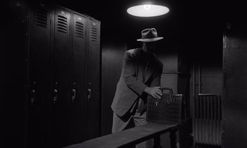 Movie still from “The Killing” (1956), directed by Stanley Kubrick – A man in a suit and hat is holding a bag; Medium shot, Low angle