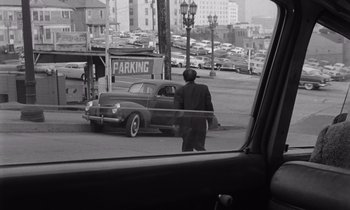 Movie still from “The Killing” (1956), directed by Stanley Kubrick – A black and white photo of a man standing in front of an old car; Wide shot, High angle
