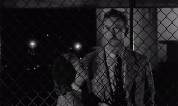 Movie still from “The Killing” (1956), directed by Stanley Kubrick – A black and white photo of a man behind a fence at night; Medium shot, Low angle