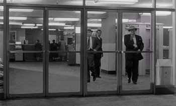 Movie still from “The Killing” (1956), directed by Stanley Kubrick – A group of men in suits and ties standing in front of a glass door; Wide shot, Low angle