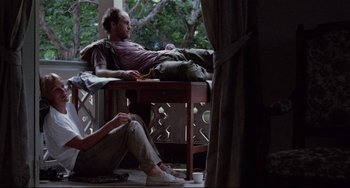 Movie still from “The Killing Fields” (1984), directed by Roland Joffé – A man sitting on top of a wooden table next to a woman; Wide shot, Low angle