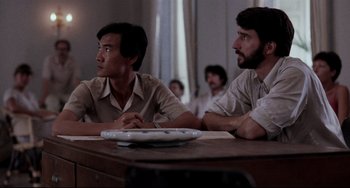 Movie still from “The Killing Fields” (1984), directed by Roland Joffé – Two men are sitting at a table and one of the men is looking on; Medium shot, Over the shoulder angle