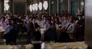 Movie still from “The Killing Fields” (1984), directed by Roland Joffé – A group of people sitting at a table in front of a bunch of lights; Wide shot, Over the shoulder angle
