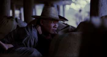 Movie still from “The Killing Fields” (1984), directed by Roland Joffé – A man wearing a straw hat is sitting on a couch; Medium shot, Over the shoulder angle