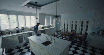 Movie still from “The Killing of a Sacred Deer” (2017), directed by Yorgos Lanthimos – A man standing on a kitchen island in front of a table; Wide shot, High angle