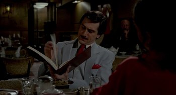 Movie still from “The King of Comedy” (1982), directed by Martin Scorsese – A man sitting at a table with a book in his hands; Medium shot, Over the shoulder angle