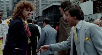 Movie still from “The King of Comedy” (1982), directed by Martin Scorsese – A man in a gray suit and a woman in a red dress are on the sidewalk; Medium shot, Over the shoulder angle