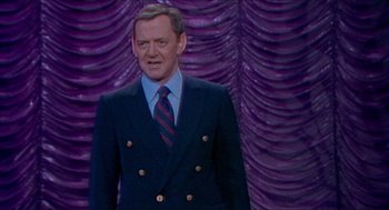 Movie still from “The King of Comedy” (1982), directed by Martin Scorsese – A man wearing a suit and tie standing in front of a purple curtain; Medium shot, Low angle