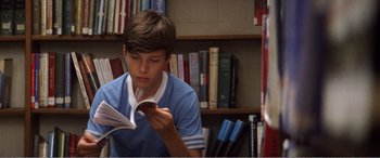 Movie still from “The Kings of Summer” (2013), directed by Jordan Vogt-Roberts – A young man reading a book in front of a bookcase; Medium shot, High angle
