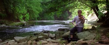 Movie still from “The Kings of Summer” (2013), directed by Jordan Vogt-Roberts – A man sitting on a rock near a river; Wide shot, Low angle