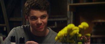 Movie still from “The Kings of Summer” (2013), directed by Jordan Vogt-Roberts – A young man smiling at the camera while holding a yellow flower; Close Up shot, Over the shoulder angle