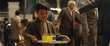 Movie still from “The Kite Runner” (2007), directed by Marc Forster – A young boy holding a tray with a glass of orange juice on top of it; Medium shot, Over the shoulder angle