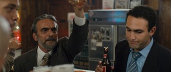 Movie still from “The Kite Runner” (2007), directed by Marc Forster – A man raising a glass at a table with a beer; Medium shot, Low angle