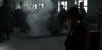 Movie still from “The Last Emperor” (1987), directed by Bernardo Bertolucci – A man wearing a hat and glasses in front of a smoke filled room; Medium shot, Over the shoulder angle