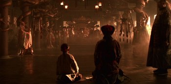 Movie still from “The Last Emperor” (1987), directed by Bernardo Bertolucci – Two people sitting on the ground in front of an audience; Wide shot, High angle