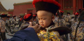 Movie still from “The Last Emperor” (1987), directed by Bernardo Bertolucci – A young boy wearing a hat and a necklace is being held up by an older person; Close Up shot, Over the shoulder angle