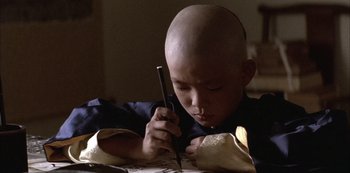 Movie still from “The Last Emperor” (1987), directed by Bernardo Bertolucci – A bald boy is holding a pen and looking down at a piece of paper on a table; Close Up shot, High angle