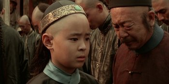 Movie still from “The Last Emperor” (1987), directed by Bernardo Bertolucci – A young boy with an oriental headdress is standing in front of a group of people; Close Up shot, Over the shoulder angle