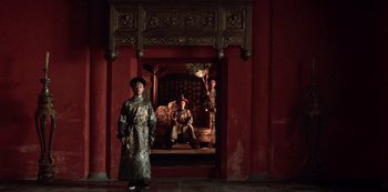 Movie still from “The Last Emperor” (1987), directed by Bernardo Bertolucci – A man standing in front of a doorway with a man sitting on a bench in the background; Wide shot, Low angle
