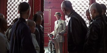 Movie still from “The Last Emperor” (1987), directed by Bernardo Bertolucci – An old man wearing a suit and a hat is holding a bicycle; Wide shot, Over the shoulder angle