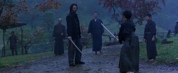 Movie still from “The Last Samurai” (2003), directed by Edward Zwick – A group of people standing in the dirt holding wooden sticks; Wide shot, Over the shoulder angle