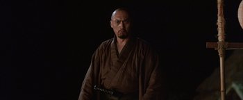 Movie still from “The Last Samurai” (2003), directed by Edward Zwick – A man in a brown robe holding a sword in front of a black background; Close Up shot, Low angle