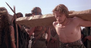 Movie still from “The Last Temptation of Christ” (1988), directed by Martin Scorsese – A man carries a wooden cross on his back; Medium shot, Low angle