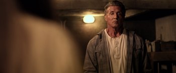 Movie still from “Rambo: Last Blood” (2019), directed by Adrian Grunberg – A man standing in a dimly - lit room looking at the camera; Close Up shot, Over the shoulder angle