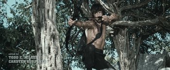 Movie still from “Rambo: Last Blood” (2019), directed by Adrian Grunberg – A shirtless man is holding a bow and arrow in the woods; Medium shot, Low angle