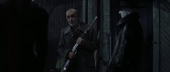 Movie still from “The League of Extraordinary Gentlemen” (2003), directed by Stephen Norrington – An older man holding a rifle in a dark room; Medium shot, Over the shoulder angle
