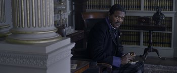 Movie still from “The Legend of Tarzan” (2016), directed by David Yates – A man in a suit sitting in a chair in front of a book case; Medium shot, High angle