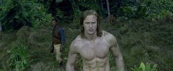 Movie still from “The Legend of Tarzan” (2016), directed by David Yates – A shirtless man in a field with a man standing behind him; Medium shot, Low angle