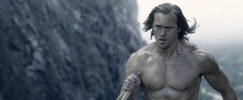 Movie still from “The Legend of Tarzan” (2016), directed by David Yates – A shirtless man holding a sword in front of a cliff; Medium shot, Over the shoulder angle