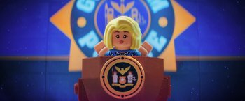 Movie still from “The Lego Batman Movie” (2017), directed by Chris McKay – A lego figure of a woman sitting at a podium in front of a clock; Medium shot, Over the shoulder angle