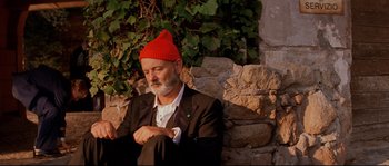 Movie still from “The Life Aquatic with Steve Zissou” (2004), directed by Wes Anderson – An older man wearing a red hat sitting against a stone wall; Medium shot, Low angle