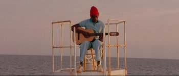 Movie still from “The Life Aquatic with Steve Zissou” (2004), directed by Wes Anderson – A man sitting on top of a chair playing a guitar in front of the ocean; Wide shot, Low angle