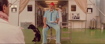 Movie still from “The Life Aquatic with Steve Zissou” (2004), directed by Wes Anderson – A man in a red hat sitting on a stool with a black dog; Wide shot, Low angle