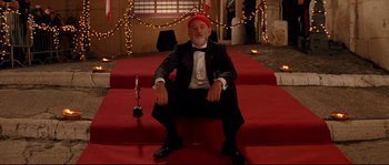 Movie still from “The Life Aquatic with Steve Zissou” (2004), directed by Wes Anderson – A man sitting on a red carpet wearing a red hat; Wide shot, Low angle