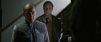Movie still from “The Lives of Others” (2006), directed by Florian Henckel von Donnersmarck – Two men standing next to each other in front of a staircase; Medium shot, Low angle