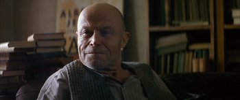 Movie still from “The Lives of Others” (2006), directed by Florian Henckel von Donnersmarck – A man with a shaved head sitting in front of a bookshelf; Close Up shot, Low angle