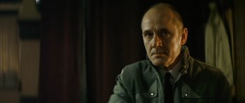Movie still from “The Lives of Others” (2006), directed by Florian Henckel von Donnersmarck – A man in a suit and tie sitting in front of a curtain; Close Up shot, Low angle
