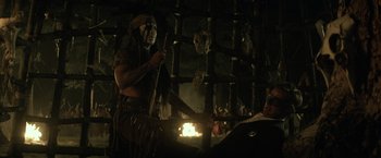 Movie still from “The Lone Ranger” (2013), directed by Gore Verbinski – A man in a native american costume holding a stick in front of a fire pit; Medium shot, Over the shoulder angle