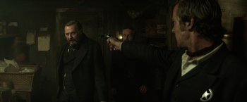 Movie still from “The Lone Ranger” (2013), directed by Gore Verbinski – A man holding a gun pointed at another man in a dark room; Medium shot, Over the shoulder angle
