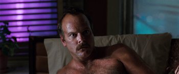 Movie still from “The Long Kiss Goodnight” (1996), directed by Renny Harlin – A man with a beard and mustache is sitting on a couch; Close Up shot, Low angle