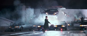 Movie still from “The Long Kiss Goodnight” (1996), directed by Renny Harlin – A man in a black coat is standing in front of a car; Wide shot, High angle