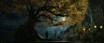 Movie still from “The Lord of the Rings: The Rings of Power” (2022), created by Patrick McKay – Two people are standing under a large tree; Extreme Wide shot, Low angle