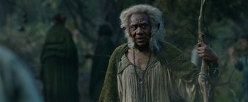 Movie still from “The Lord of the Rings: The Rings of Power” (2022), created by Patrick McKay – A man with a long beard and a long necklace is standing in the woods; Close Up shot, Low angle