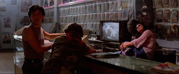 Movie still from “The Lost Boys” (1987), directed by Joel Schumacher – A man sitting at a table in front of a television; Medium shot, Over the shoulder angle