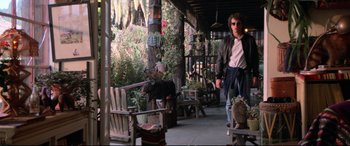 Movie still from “The Lost Boys” (1987), directed by Joel Schumacher – A man walking down a porch next to wooden chairs; Wide shot, Over the shoulder angle
